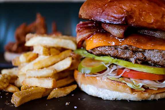 A burger with bacon, cheese, tomato, and pickles on a bun with fries on the side.