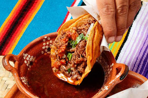 A person is holding a taco with meat and vegetables in a clay dish.