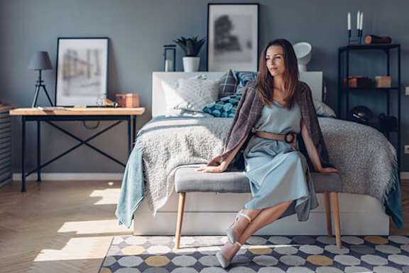 A woman in a grey dress and brown cardigan is sitting on a bed with a grey blanket.