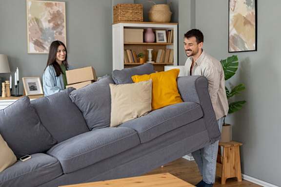 A man and woman are moving a grey couch into a room.