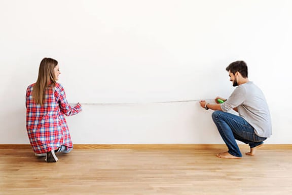 A man and a woman are sitting on the floor, facing a wall, with a measuring tape between them.