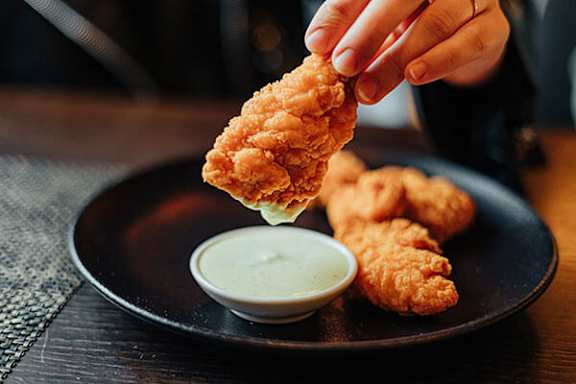 A person is dipping a piece of fried chicken into a bowl of sauce.