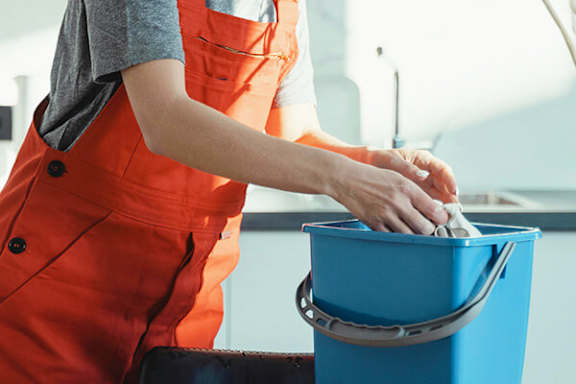 A person in an orange jumpsuit is cleaning a blue bucket with a cloth.