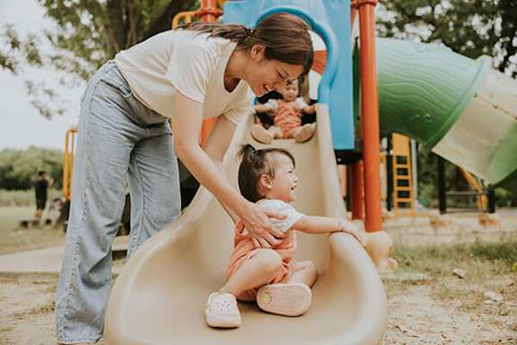 A woman helps a child slide down a slide at a playground.