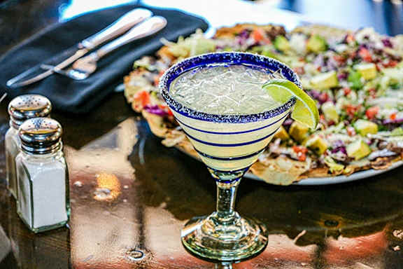 A margarita glass with a salted rim sits on a table next to a plate of nachos.