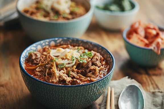 A bowl of noodles with shredded meat and garnish on a wooden table.