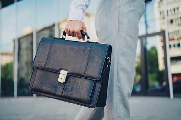 A man in a white shirt and grey pants is holding a black briefcase.