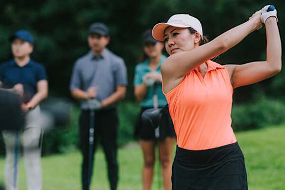 A woman in an orange top and black skirt is swinging a golf club.