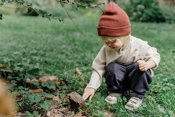 A child in a red hat is playing with a rock in the grass.