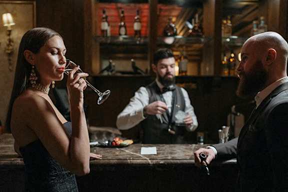 A woman in a black dress is drinking from a wine glass in a bar.