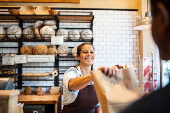 A woman in an apron is holding a bag of bread in a bakery.