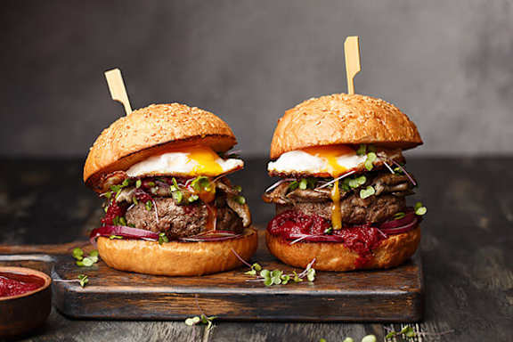 Two gourmet burgers with beef, cheese, and vegetables on a wooden board.