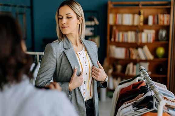 A woman in a grey blazer is talking to another person in a room with a bookshelf.