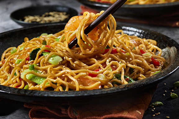 A black bowl filled with noodles and vegetables with a pair of chopsticks lifting some noodles.