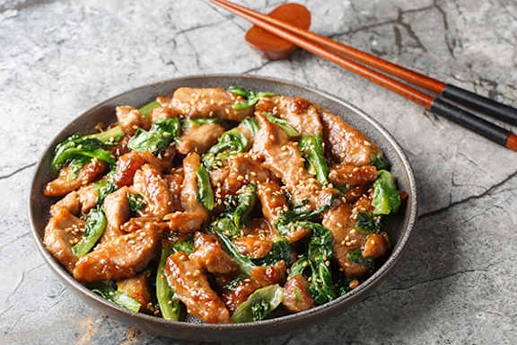 A bowl of stir-fried chicken with greens and sesame seeds, accompanied by chopsticks.
