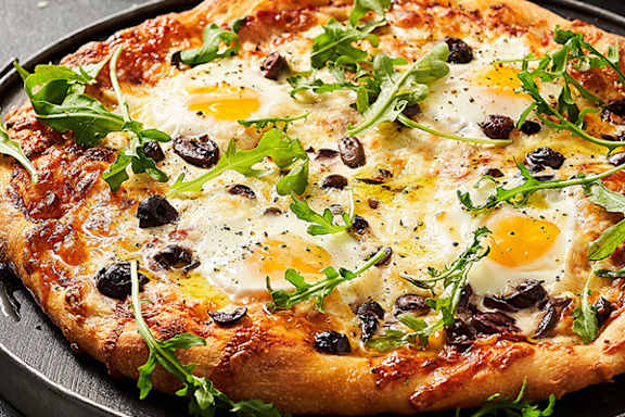 A pizza with eggs, olives and arugula on a black plate.