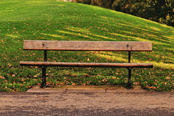 A wooden bench with metal legs sits on a concrete slab in front of a grassy hill.