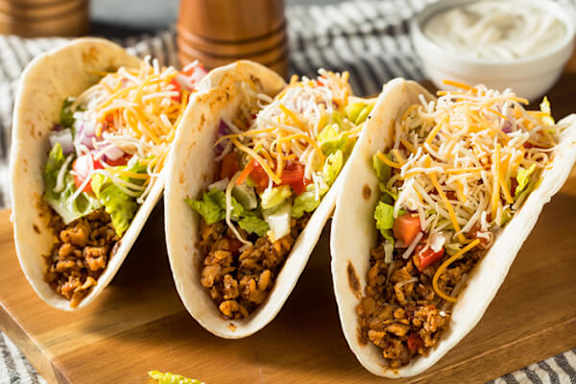 Three tacos with cheese on top are on a wooden board.