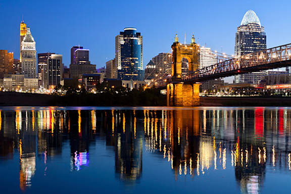 A city skyline at dusk with a bridge and buildings reflected in the water.