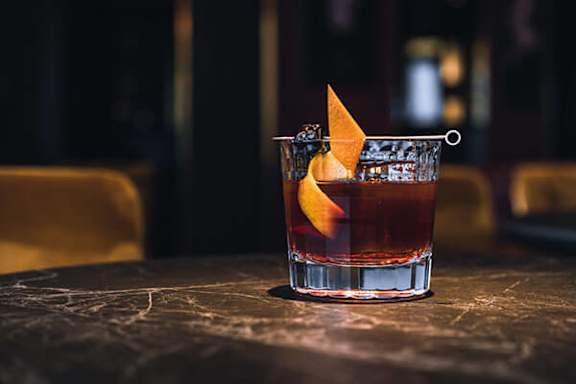 A glass of a brown drink with a lemon peel garnish on a table.