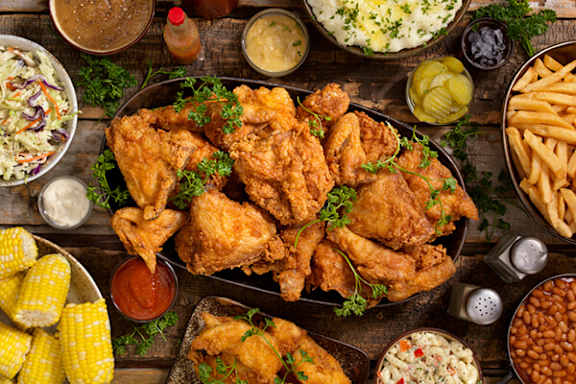 A variety of food including fried chicken, corn, beans, and coleslaw are displayed on a wooden table.