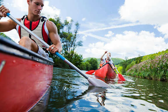 A man in a red kayak paddles down a river with another kayaker in the distance.