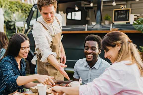 A group of people are gathered around a table, with one person serving food to the others.