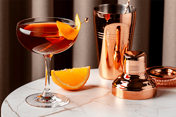 A copper-colored cocktail shaker sits next to a glass with a drink and an orange slice.