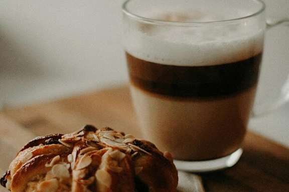 A pastry with almonds on top sits next to a glass of coffee.