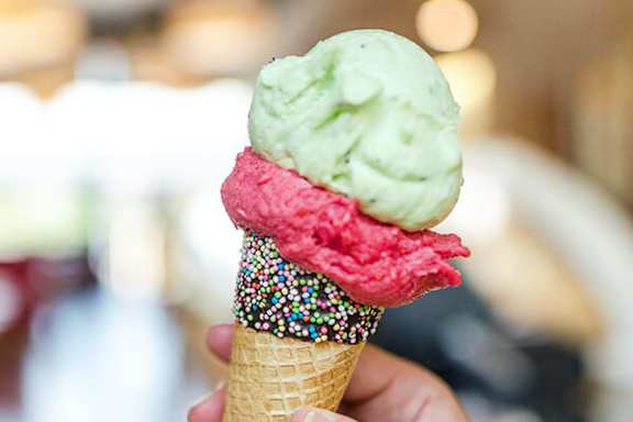 A hand is holding a cone with three scoops of ice cream in pink, green and red flavors.