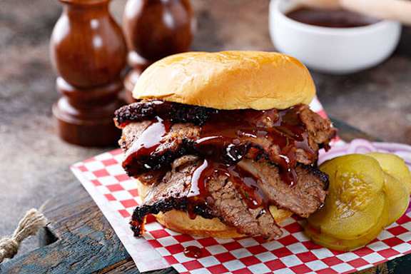A sandwich with barbecue sauce and pickles on a red and white checkered paper.