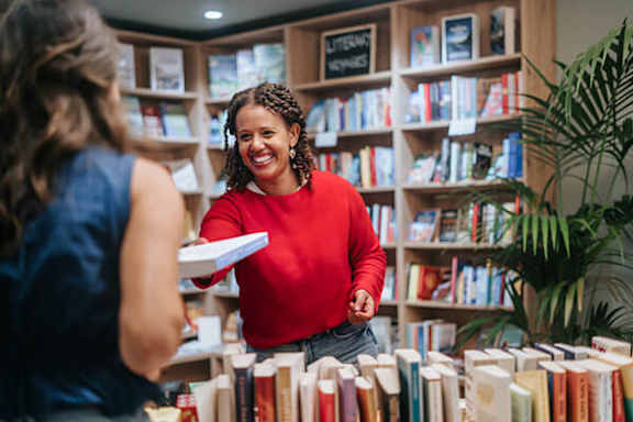 A woman in a red top is holding a book and talking to another woman in a blue top in a bookstore.