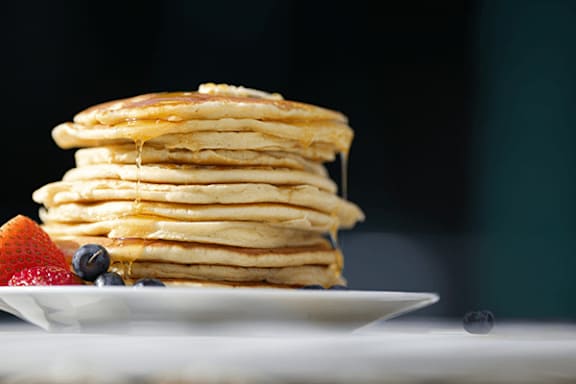 A stack of pancakes with syrup being poured on top, accompanied by a strawberry and blueberries.