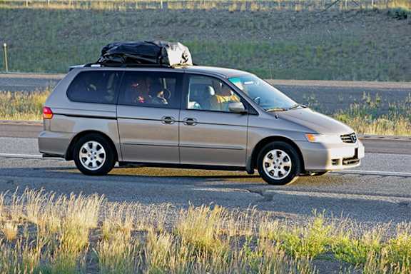 A silver minivan with a roof box and a spare wheel on the back is driving on a road.