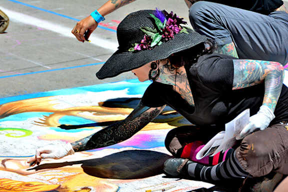 A person with a hat and tattoos is sitting on the ground drawing on the pavement.