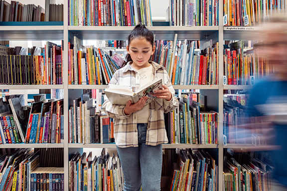 A girl is reading a book in a library with shelves full of books.