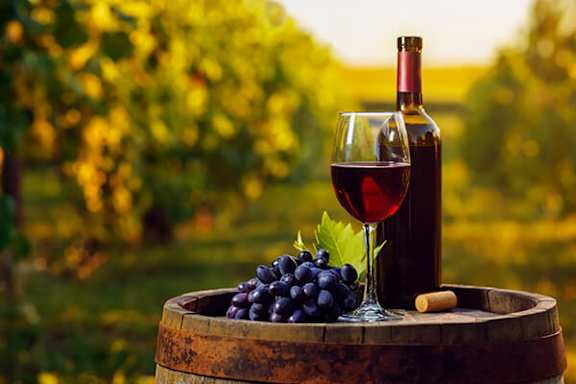 A glass of red wine and a bottle are on a barrel with a bunch of grapes.