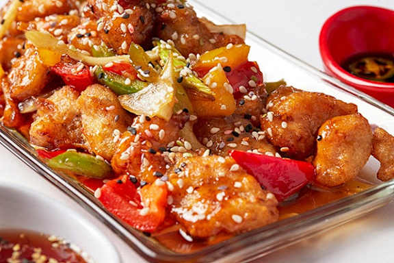 A dish of sesame chicken with vegetables and a side of dipping sauce.