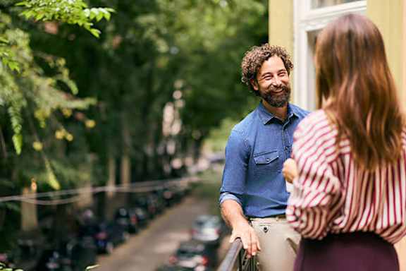 A man and a woman are standing on a balcony with a view of a street lined with parked cars.