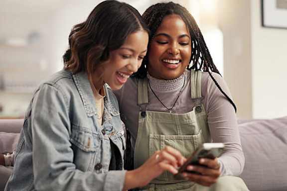Two women are sitting on a couch, one is showing something on her phone to the other.