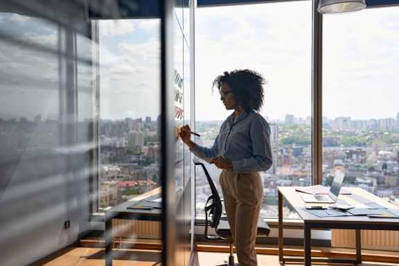A woman is writing on a whiteboard in a room with a city view.