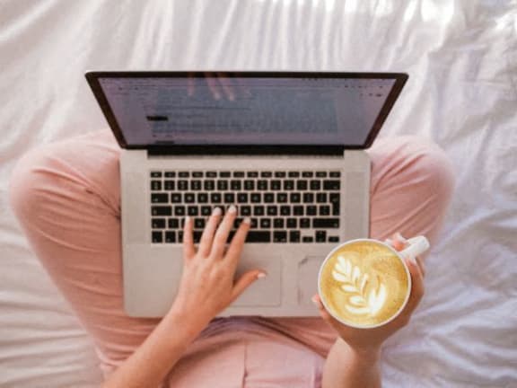 a woman sitting on a bed with a laptop and a cup of coffee