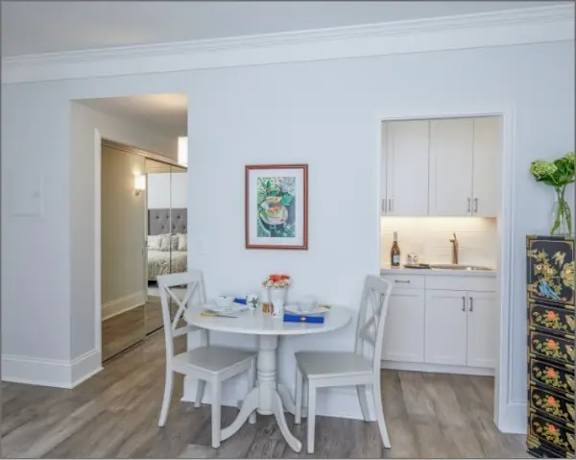 A dining room with a white table and chairs, a framed picture on the wall, and a kitchen area with white cabinets in the background.