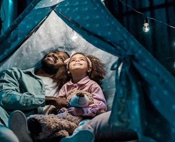 a father and daughter laying in a tent with a teddy bear