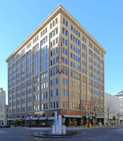 A tall building with many windows is in the middle of a plaza with a fountain.