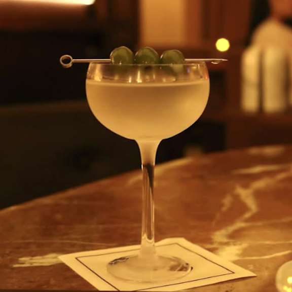 A glass of a clear drink with a green olive on the rim.