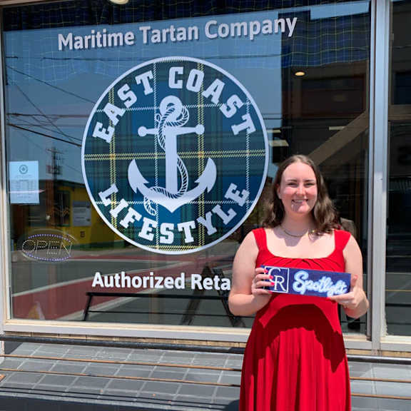 A woman in a red dress stands in front of a window with a sign that reads "Maritime Tartan Company East Coast Lifestyle."
