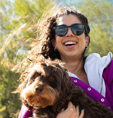 A woman in sunglasses and a purple sweater is holding a brown dog.