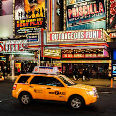 A yellow taxi is in front of a lit-up billboard for Priscilla Queen of the Desert. at The Ellington Apartments, New York, NY, 10019