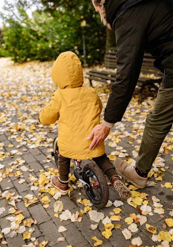 A child in a yellow jacket is learning to ride a bike.